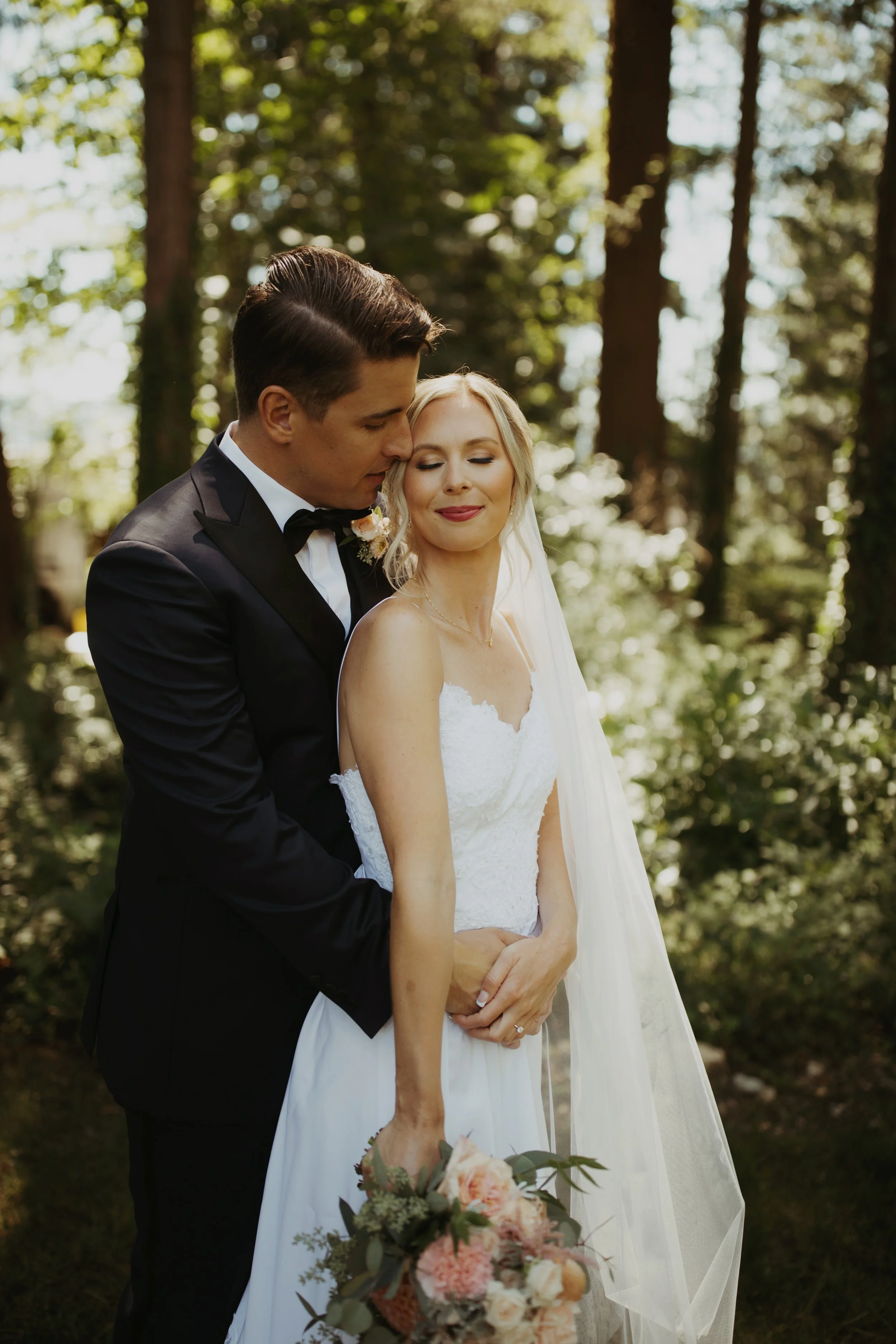 A bride and groom embrace outdoors in a wooded area during their wedding, with the groom in a black tuxedo and the bride in a white wedding dress, holding a bouquet of flowers.