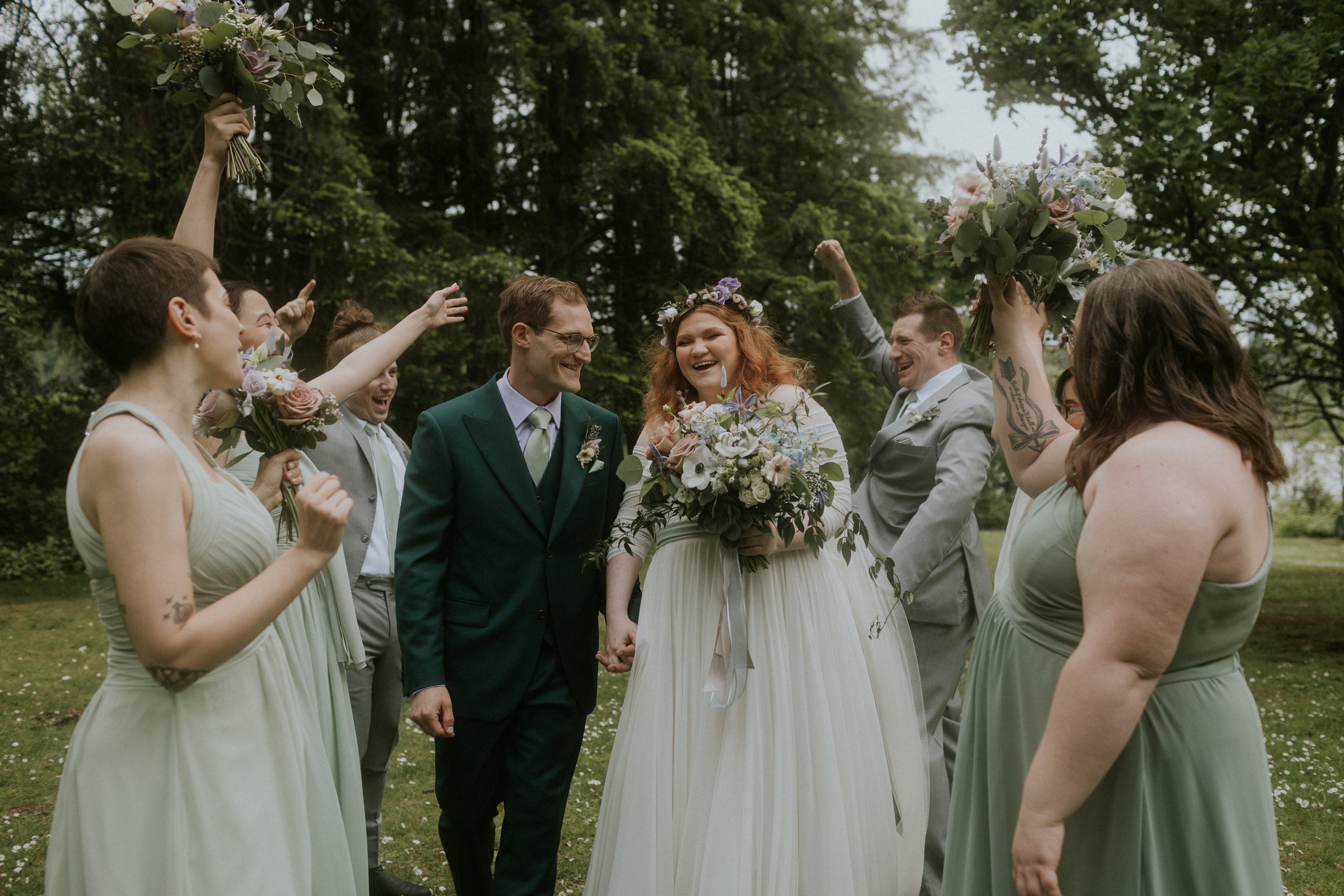 A wedding scene with a bride in a white dress holding a bouquet, standing next to a groom in a green suit. They are surrounded by happy bridesmaids and groomsmen outdoors, with all celebrating and smiling.