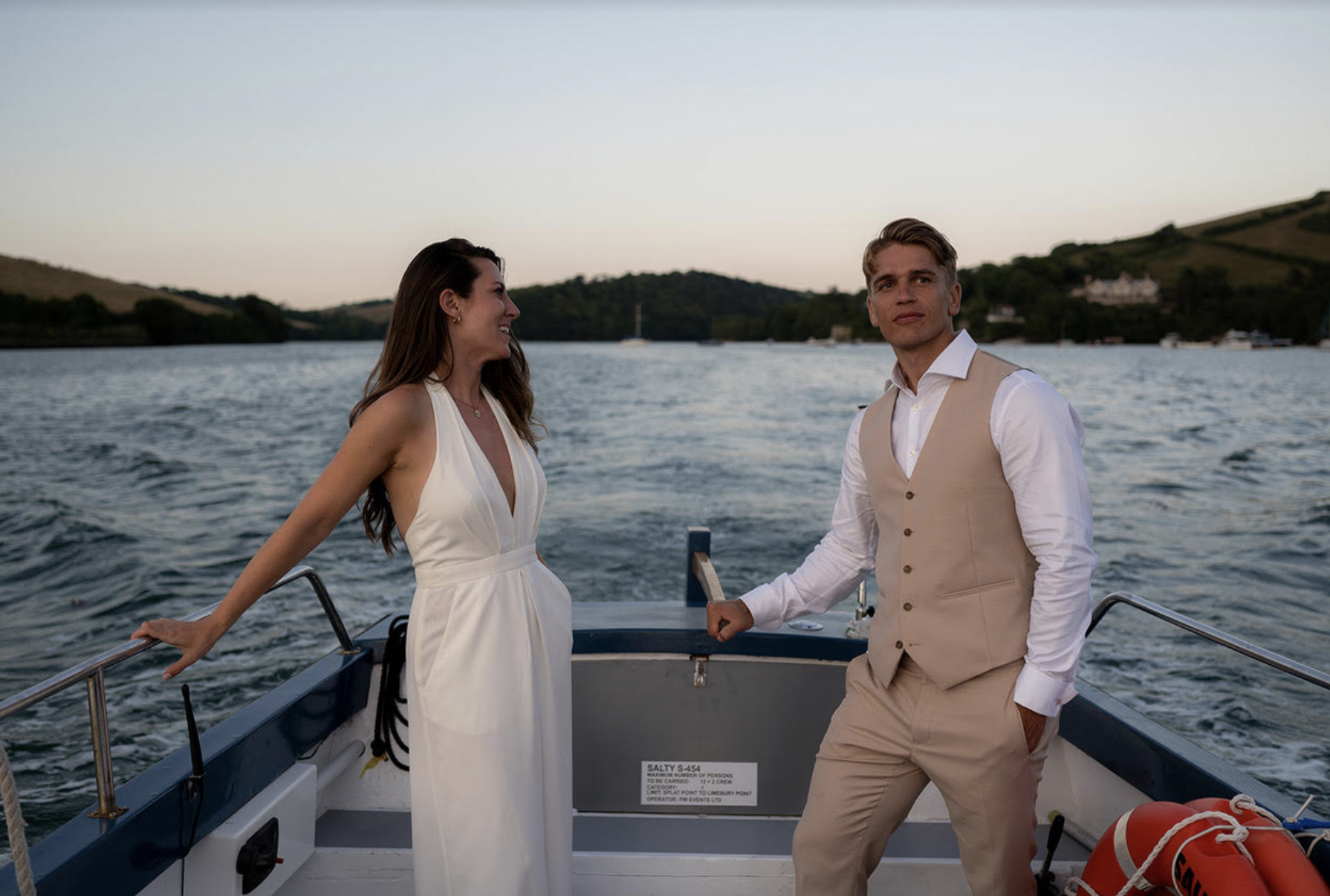 A man and woman dressed in formal attire on a boat, with the woman smiling and looking at the man, and the man looking into the distance, with water and hilly shoreline in the background.