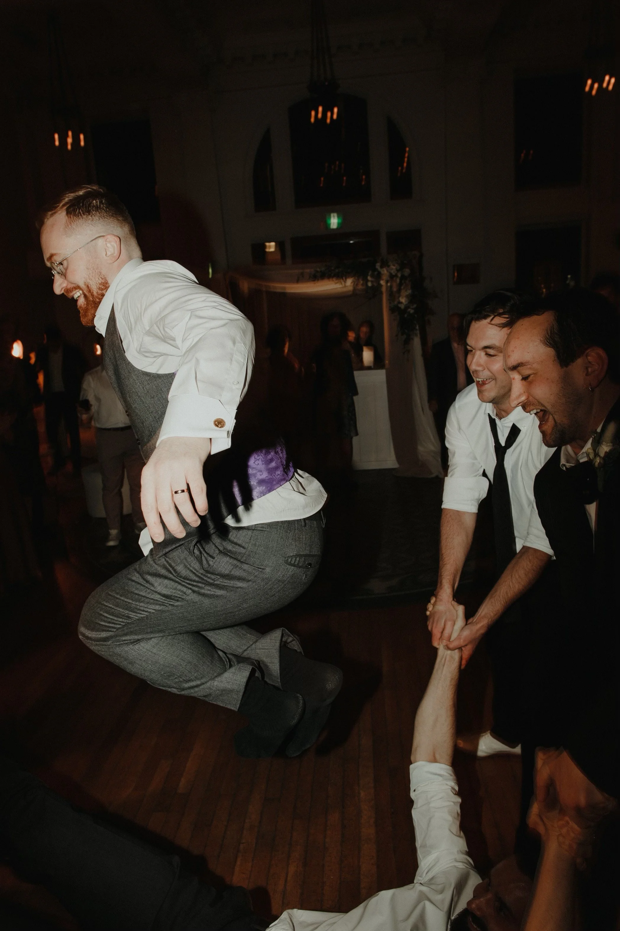 Men dancing and playing at a celebration. One man is jumping over another who is holding his hand. The scene is indoors with dim lighting and chandeliers.