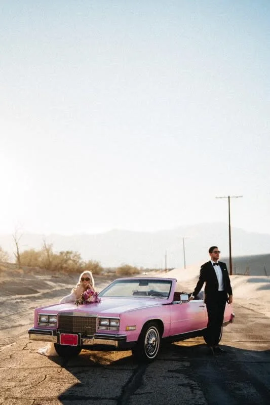 A woman and a man dressed in formal attire with sunglasses, next to a pink vintage convertible car on a deserted road in a desert landscape.