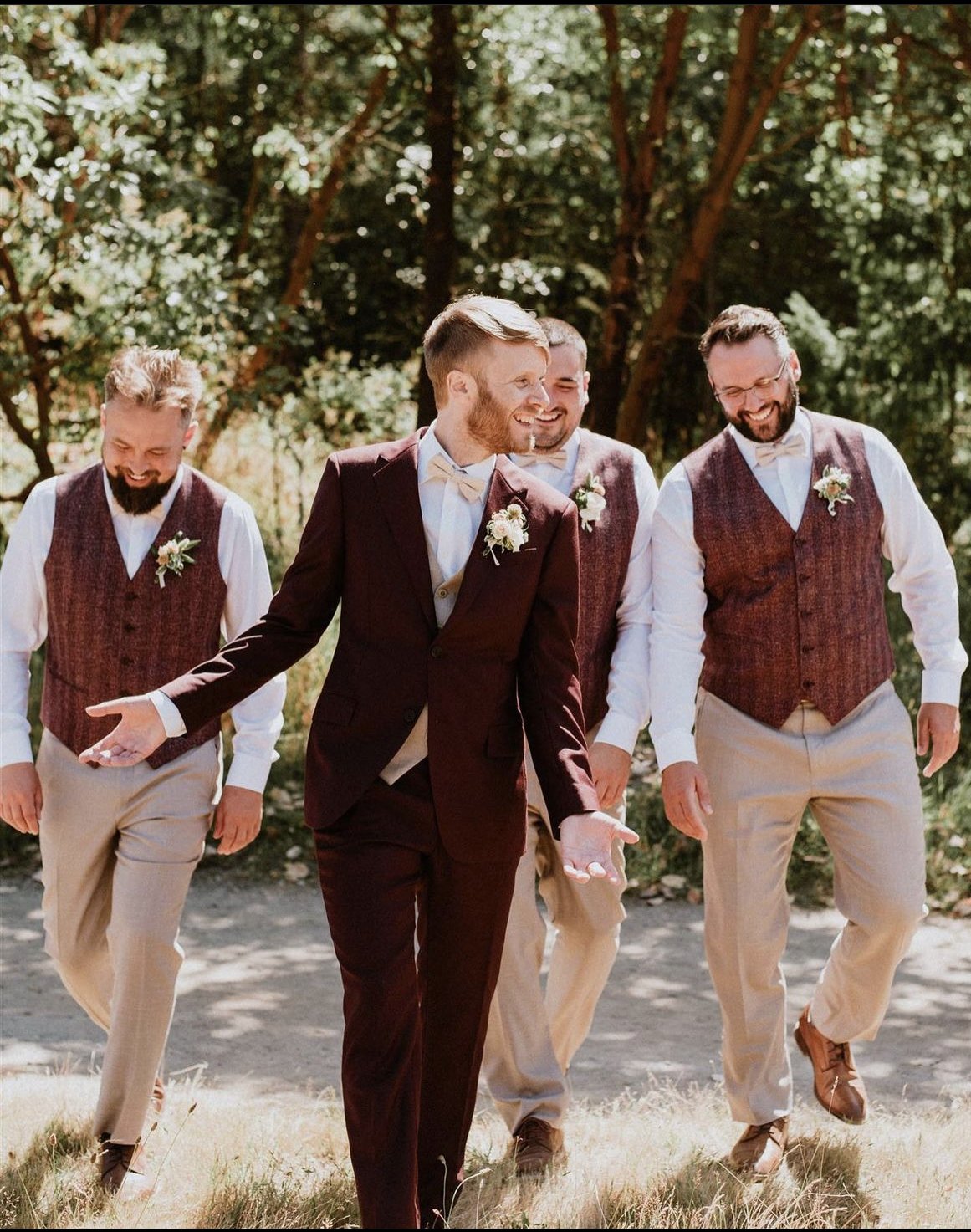 A groom in a burgundy suit walking outdoors with four groomsmen in matching vests and trousers, all smiling and enjoying a casual moment.