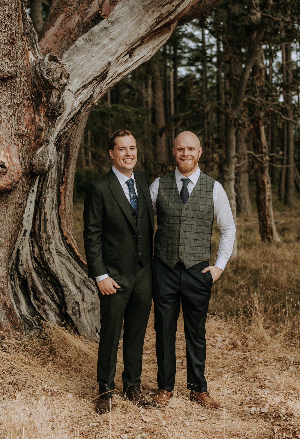 Two men in suits standing outdoors beside a large tree in a forest. One man is in a black suit with a blue tie, and the other is in a grey plaid vest with a white shirt and black tie. They are smiling.