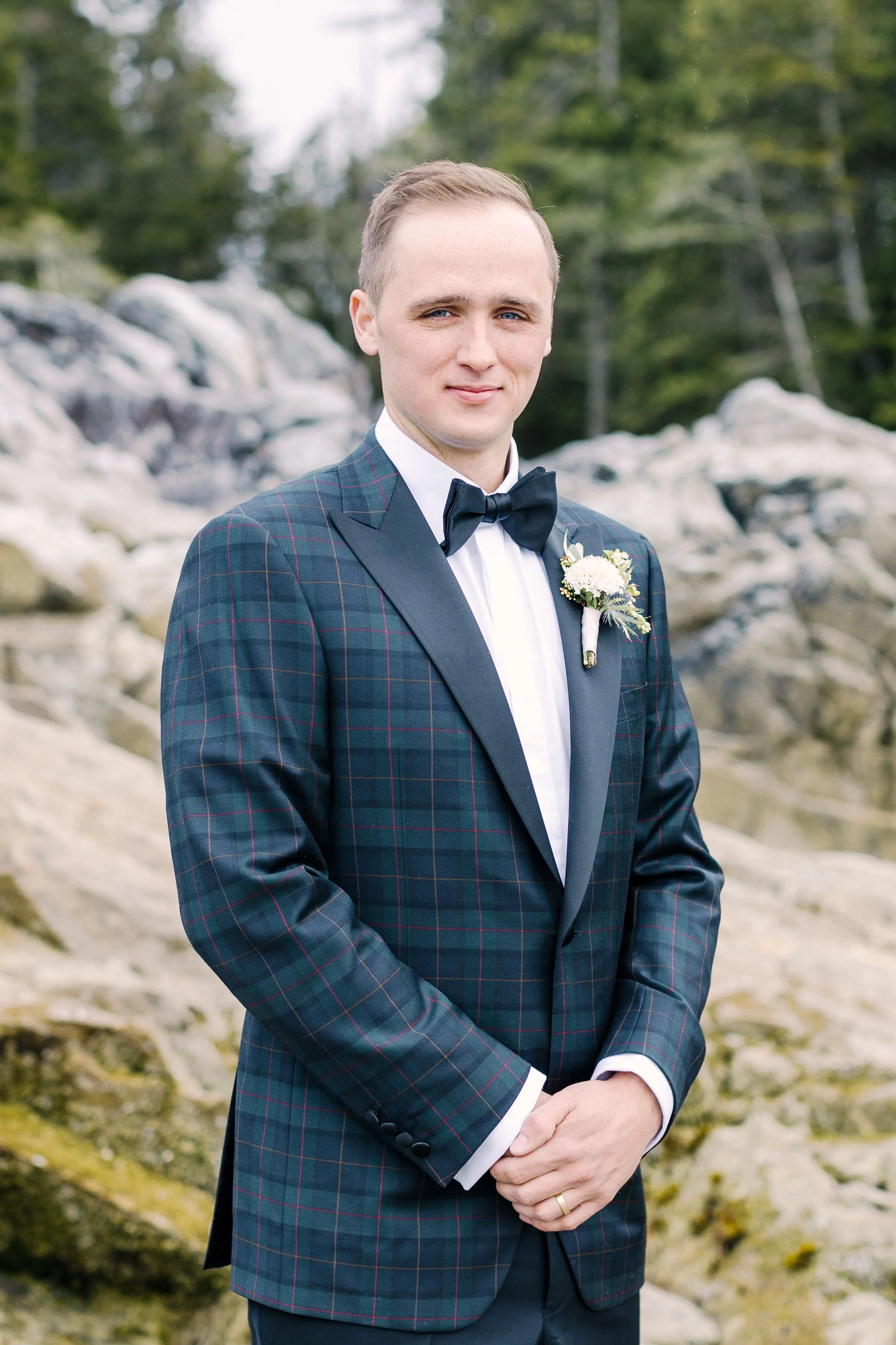 Portrait of a groom in a checked suit with a boutonniere, outdoors near rocks and greenery.