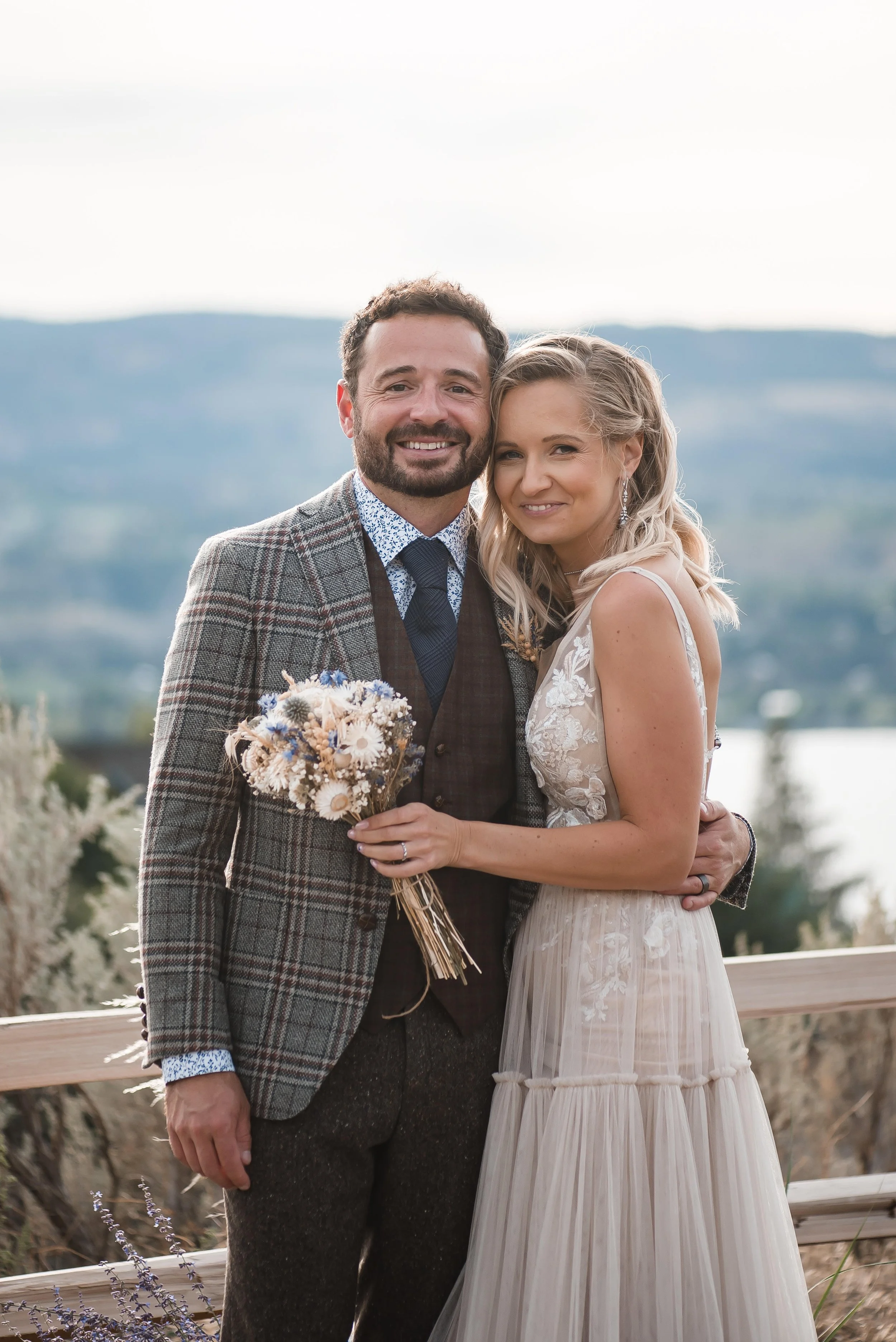 A smiling couple, a bride and groom, dressed in wedding attire, standing outdoors with mountains in the background. The bride holds a bouquet of flowers.