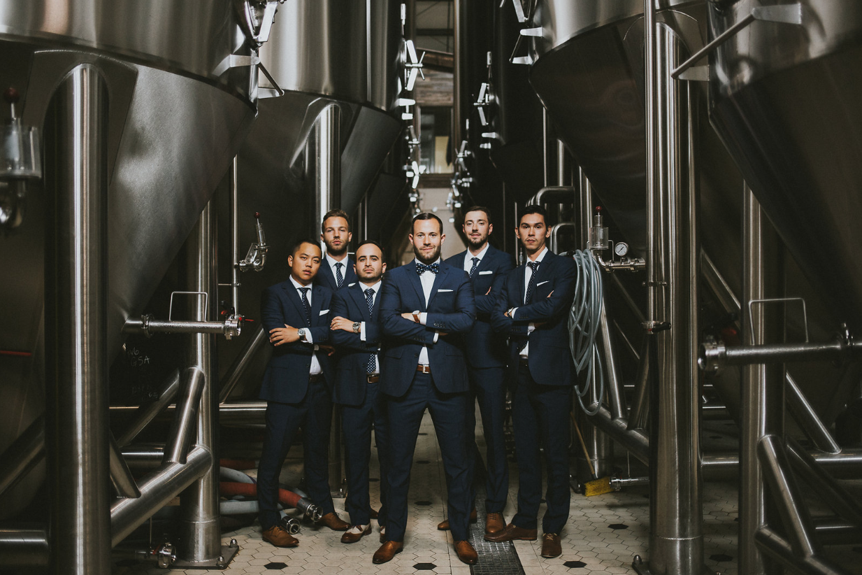 Six men in business suits standing with arms crossed in a brewery or industrial facility with large metal tanks around them.