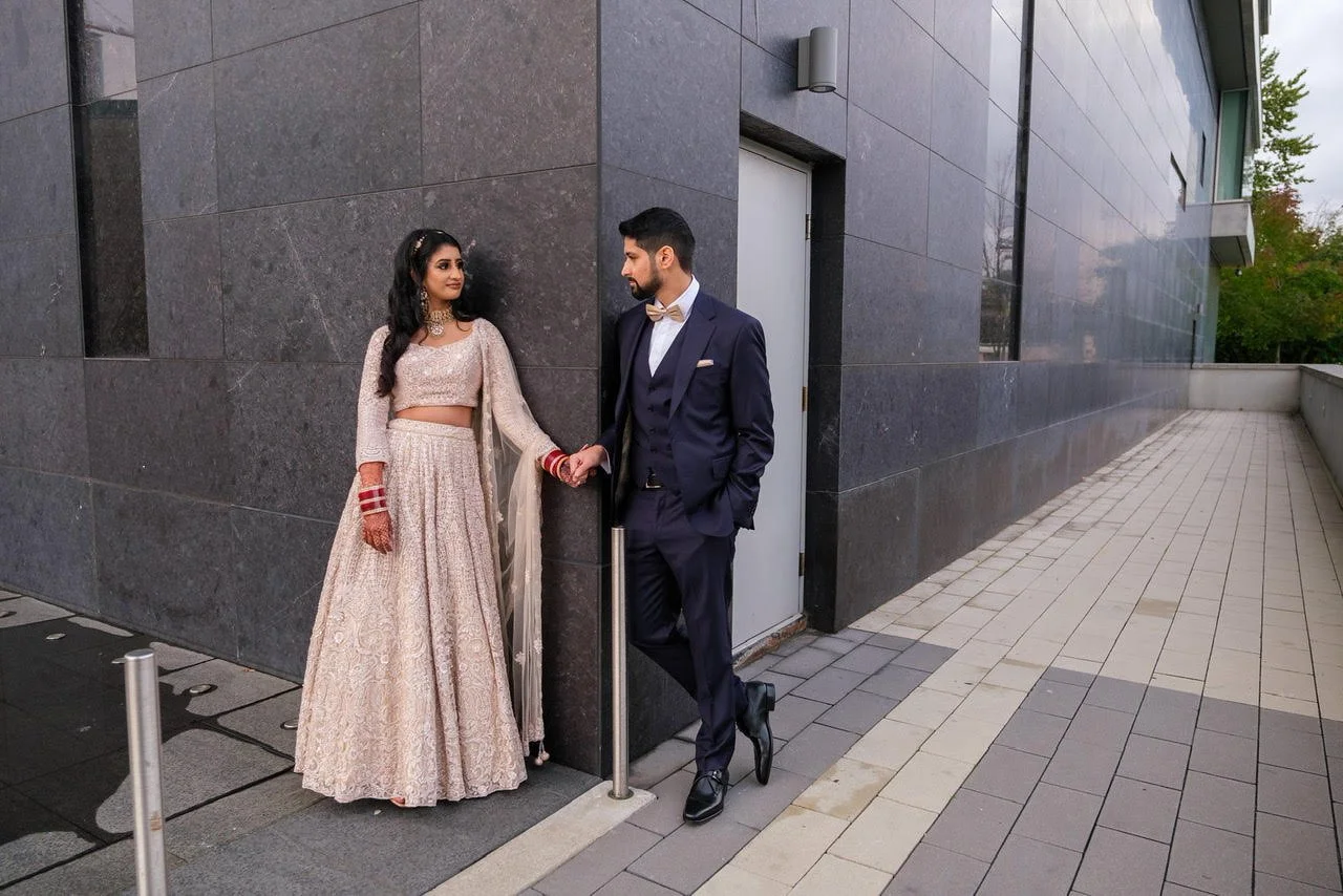 A couple dressed in traditional Indian attire standing outside a modern building. The woman wears a beige and pink Lehenga with jewelry, while the man is in a navy suit with a bow tie. They are holding hands and looking at each other.