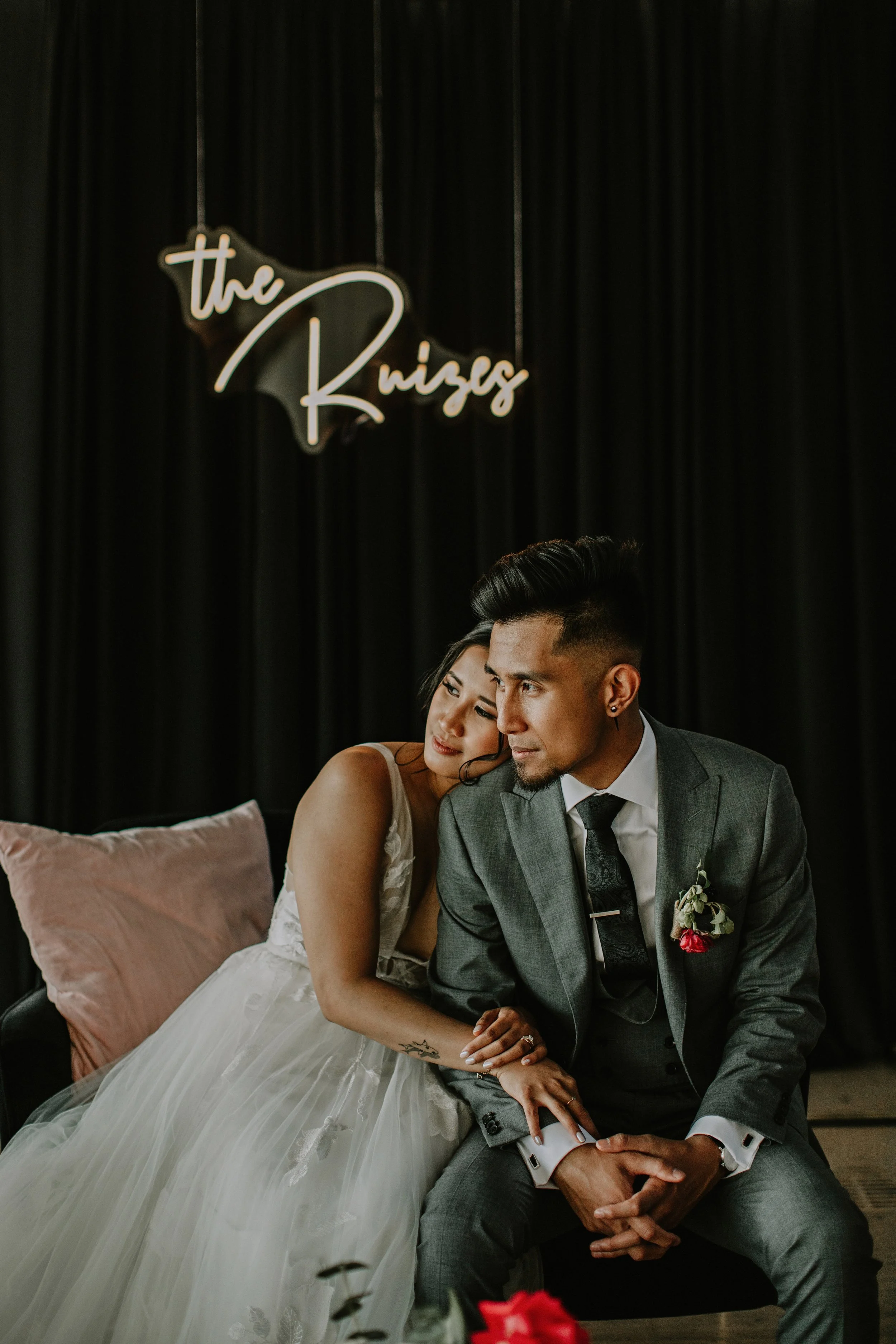 A bride and groom seated together during a wedding, with a black curtain backdrop and a neon sign reading 'the Ruizes', featuring both a man in a gray suit and white shirt and a woman in a white wedding dress.