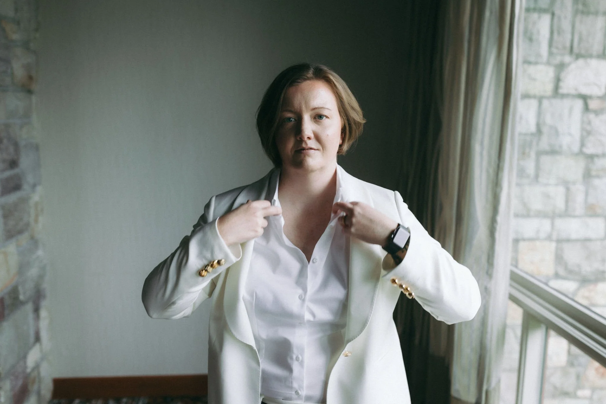 A woman with short brown hair wearing a white blazer and shirt, adjusting her blazer, standing by a window with curtains, in a room with a brick wall.