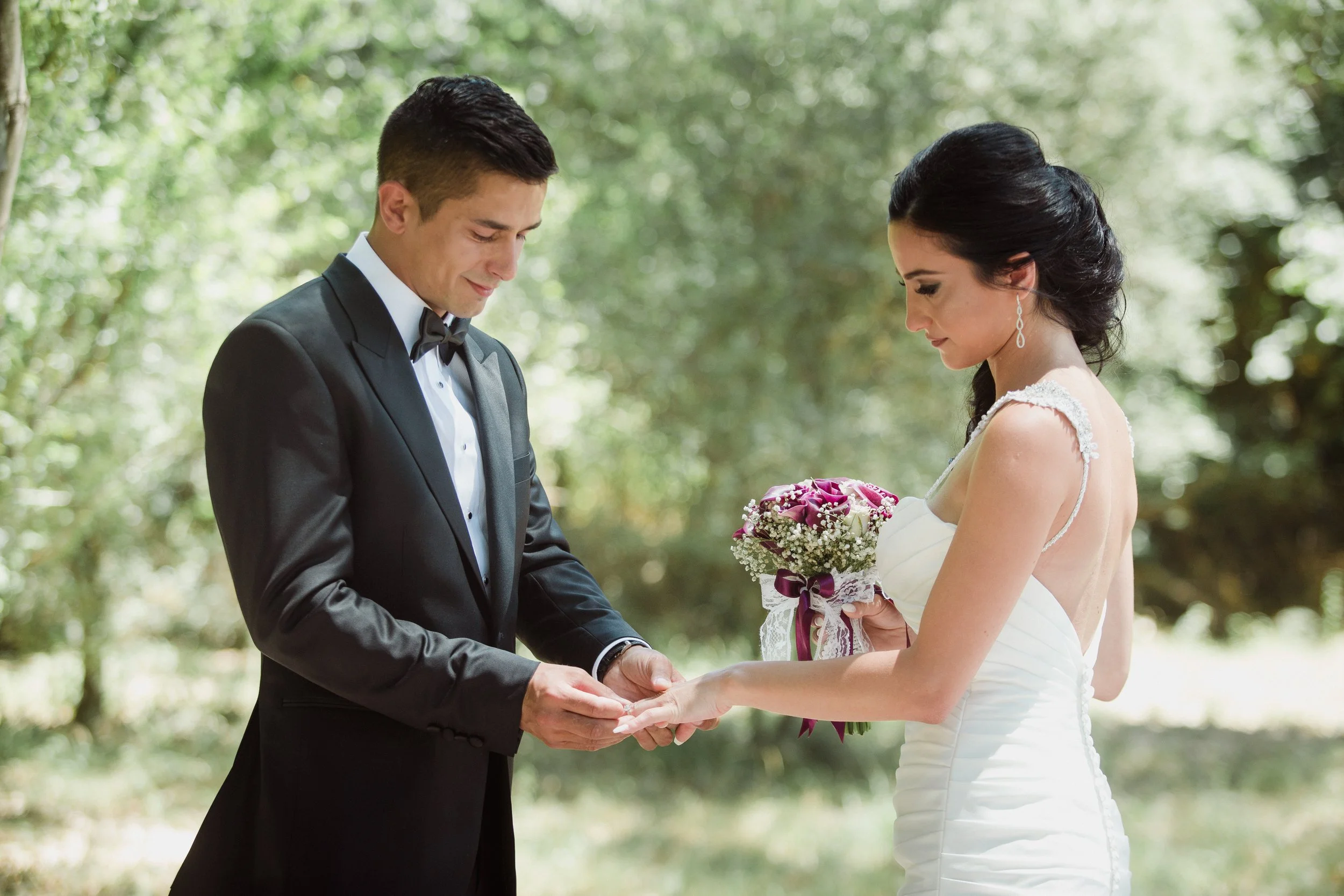 A groom dressed in a black tuxedo with a bow tie places a ring on the bride's finger during a wedding ceremony outdoors, with trees and greenery in the background.