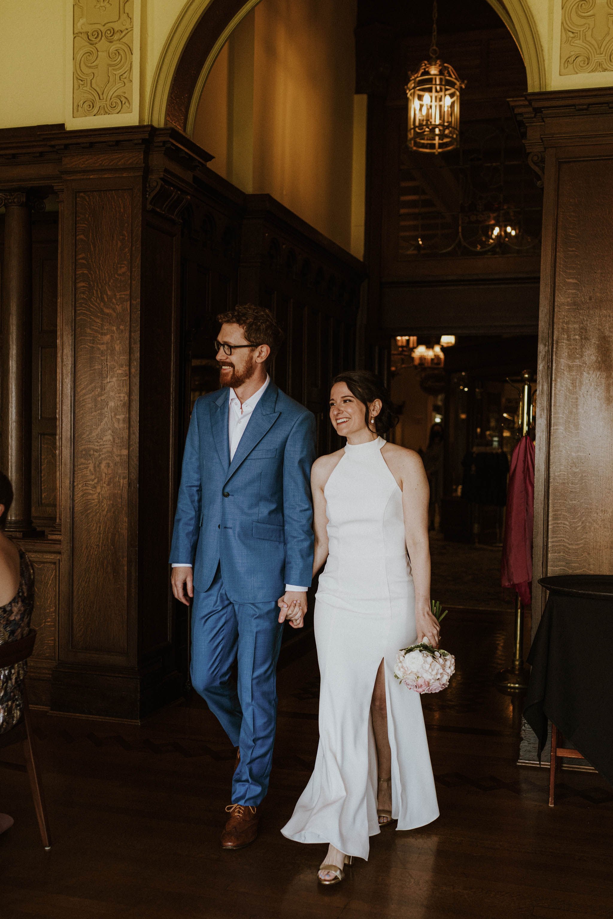 A happy bride and groom holding hands and smiling as they walk into a wedding reception. The bride is wearing a white wedding dress with a thigh-high slit and holding a bouquet, while the groom is in a blue suit and glasses. They are in a warmly lit 