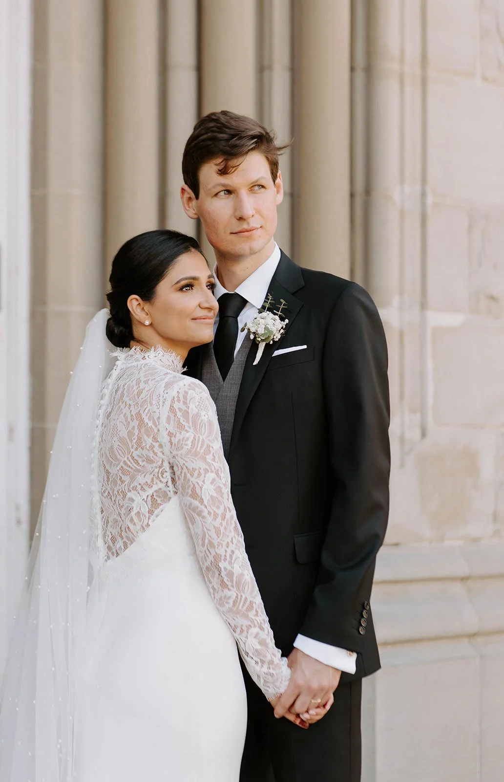 A bride and groom holding hands, standing close together outside a stone building, dressed in wedding attire, looking off to the side.