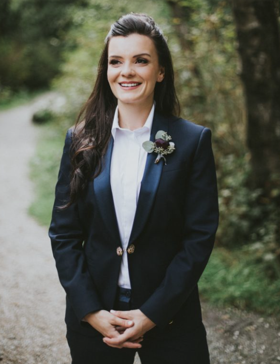 A woman in a navy blazer and white shirt standing outdoors on a path surrounded by greenery, smiling, with dark hair and a boutonniere on her lapel.