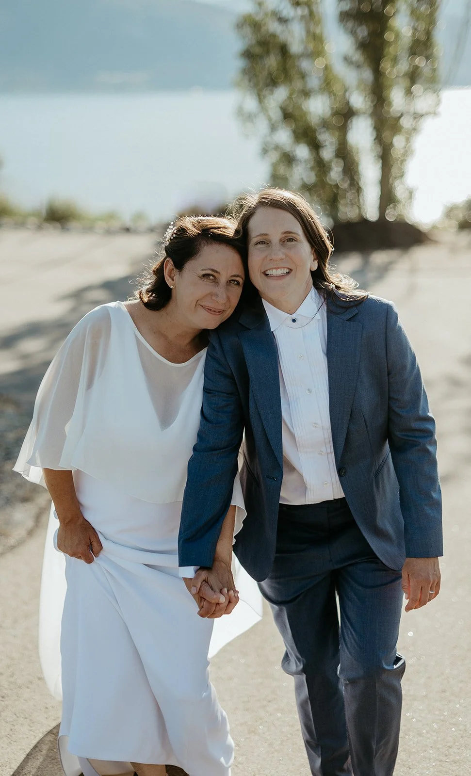 Two women, one in a white dress and the other in a dark suit, holding hands and smiling outdoors near a body of water, with a tree in the background.