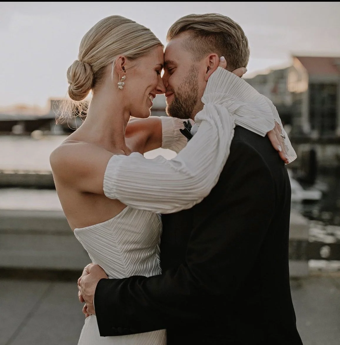 A couple dressed in wedding attire embracing each other outdoors near a waterfront at sunset.
