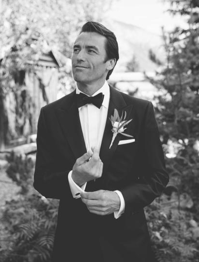 Black and white photo of a man in a tuxedo with a bow tie and boutonniere, standing outdoors with trees, smirking and holding his lapel.