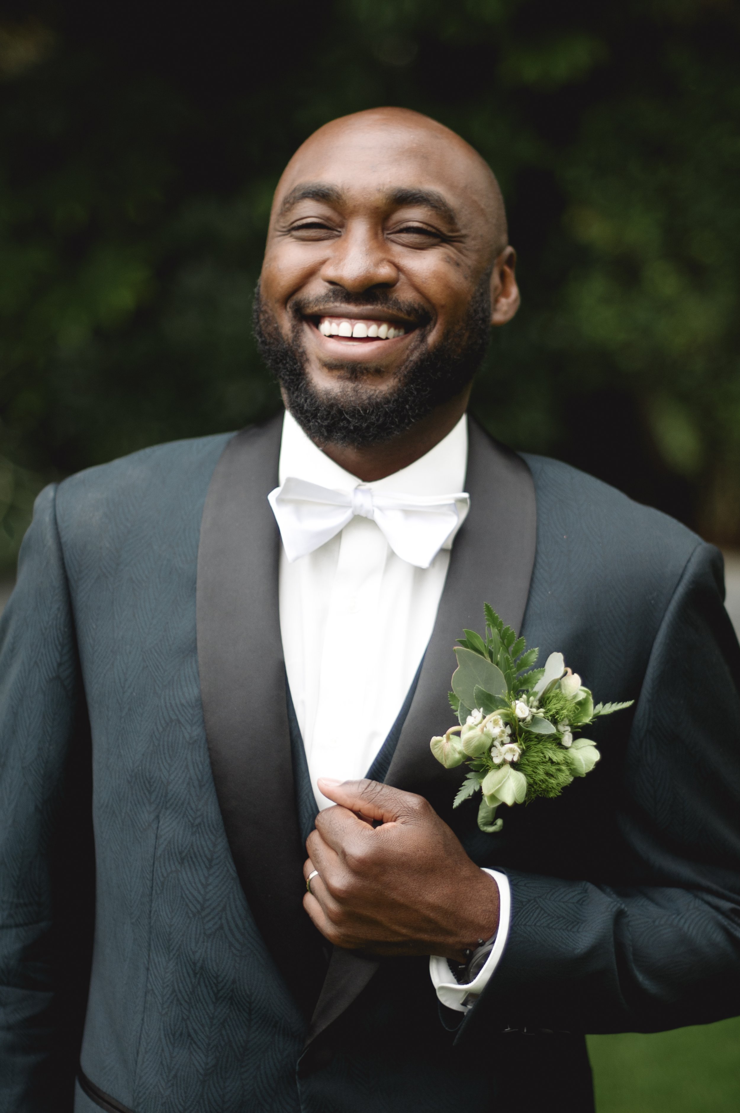 A smiling man dressed in a tuxedo with a white bow tie, holding a boutonniere with green leaves and white flowers on his lapel.