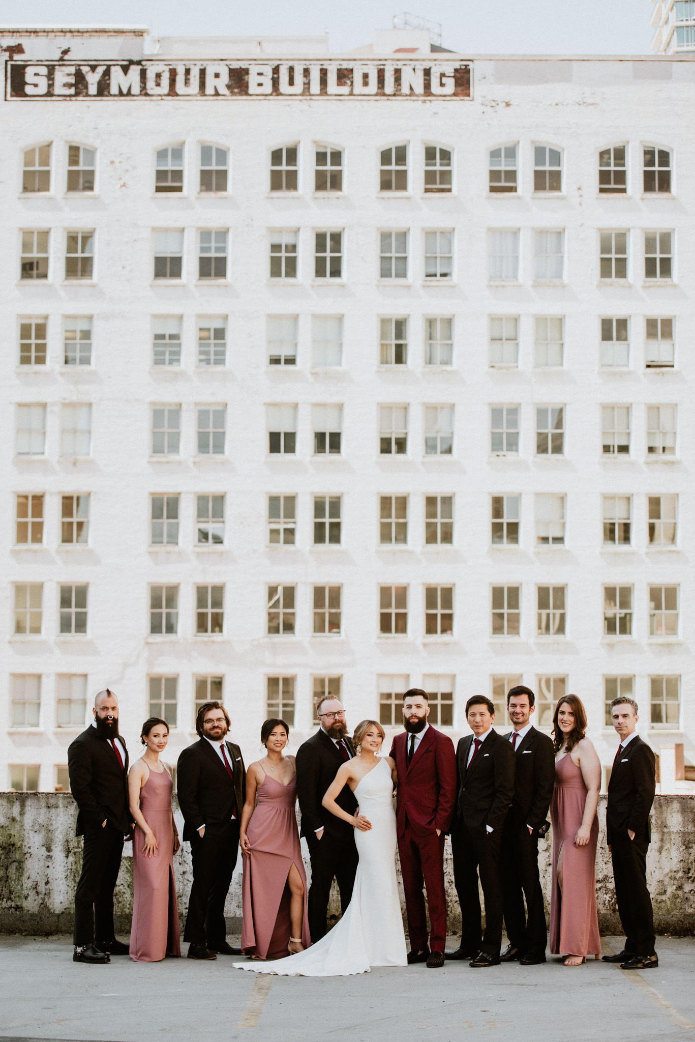 A wedding party standing outdoors in front of a building with a large sign reading 'SEYMOUR BUILDING', consisting of nine women and five men, dressed in formal attire, with the bride in a white gown and the groom in a maroon suit.