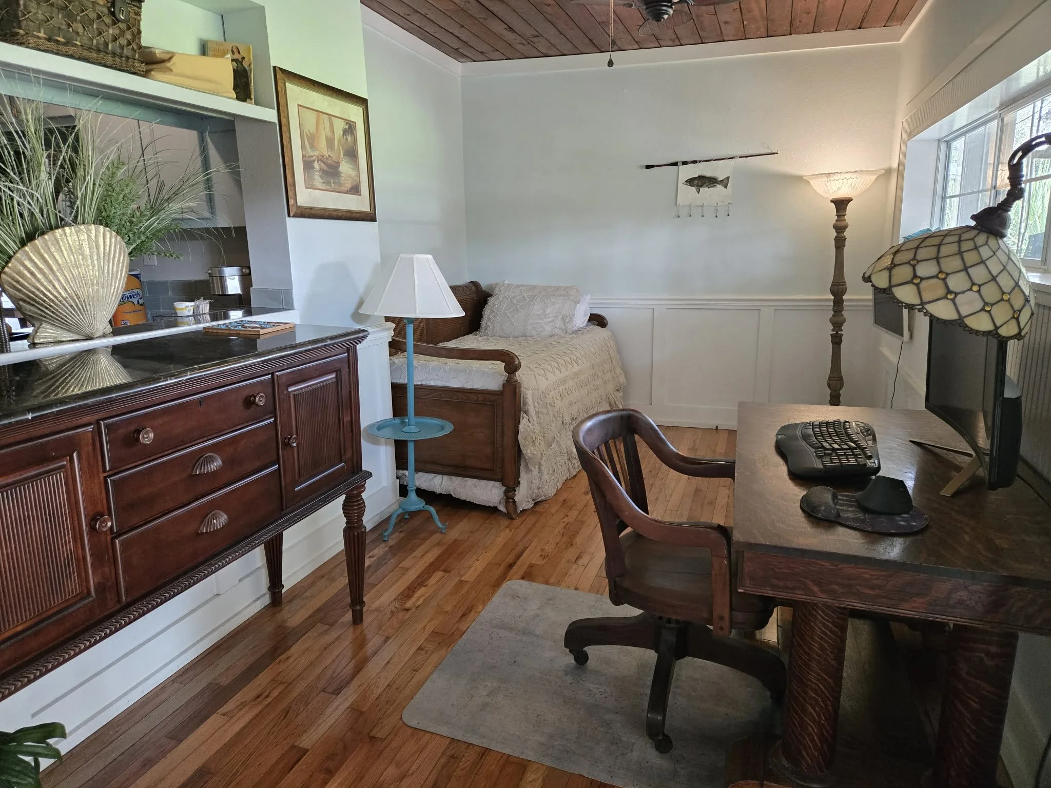 Cozy living room with a wooden sideboard, a bed with white bedding, a floor lamp, a desk with a computer, and large windows.