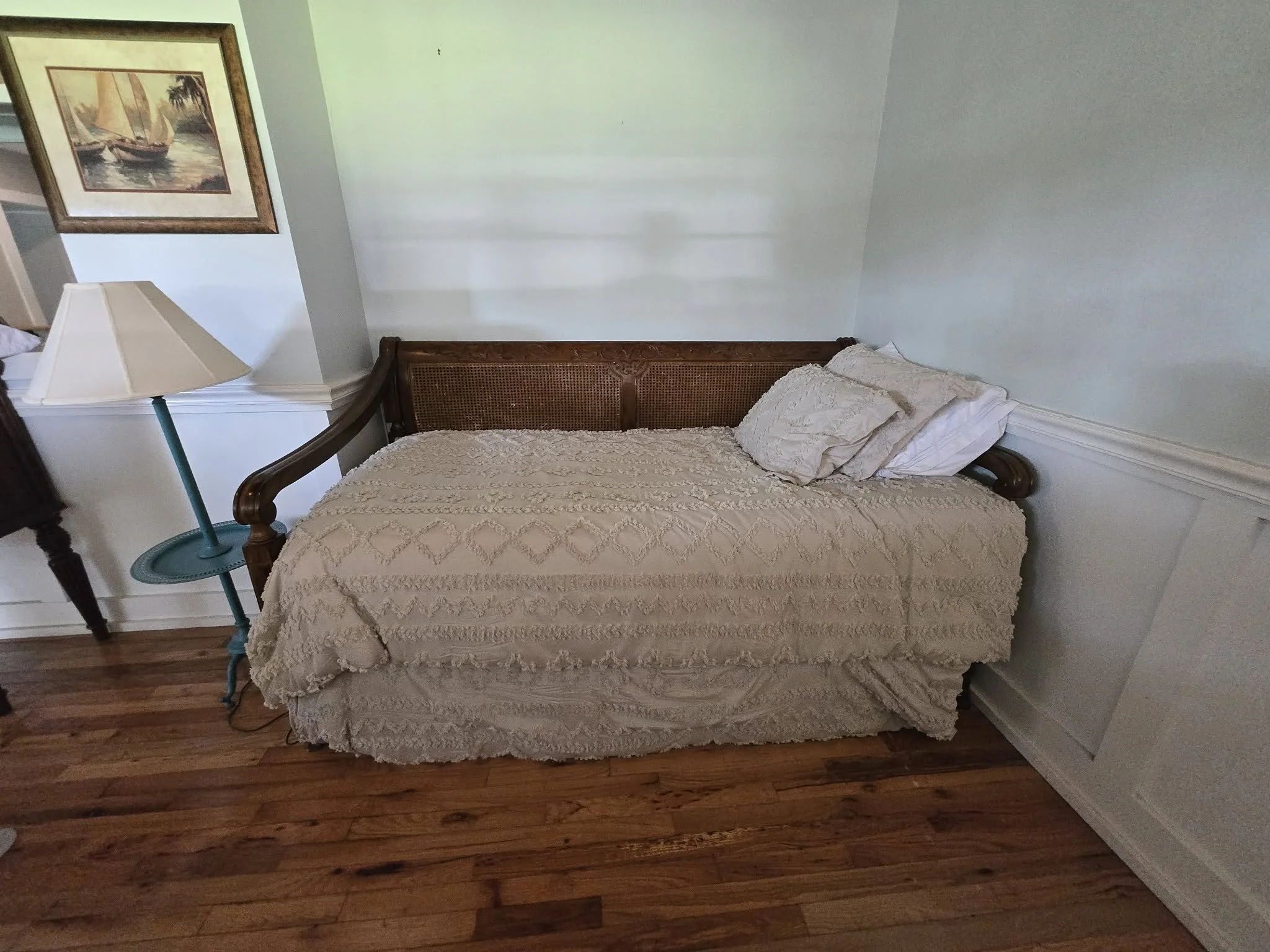 A bedroom with a wooden bed frame and a textured beige bedspread. There are two pillows at the head of the bed. To the left, a turquoise lamp and a framed painting of sailboats on water hang on the wall.