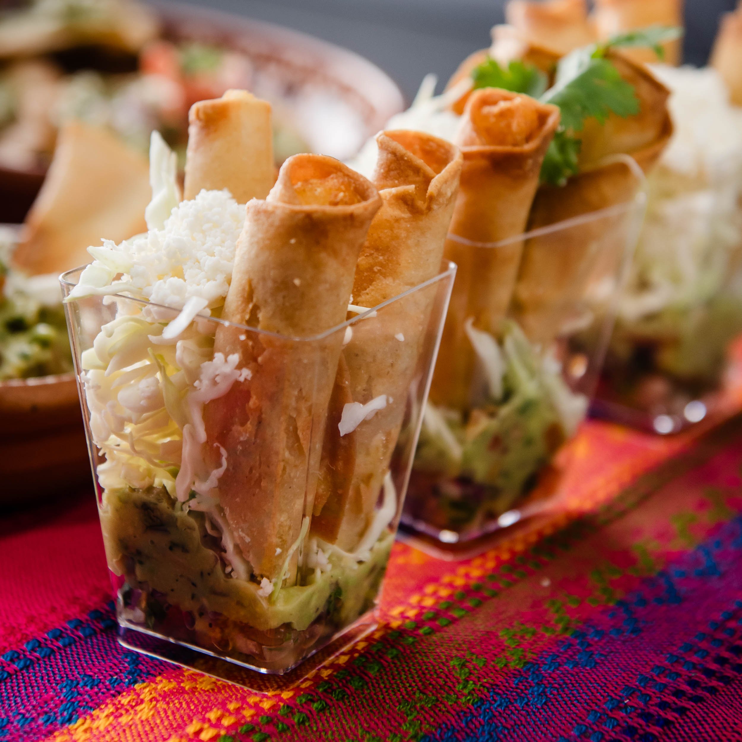 Close-up of layered Mexican appetizers in clear glasses, featuring crispy taquitos, shredded cheese, lettuce, and other toppings, on a colorful woven tablecloth.