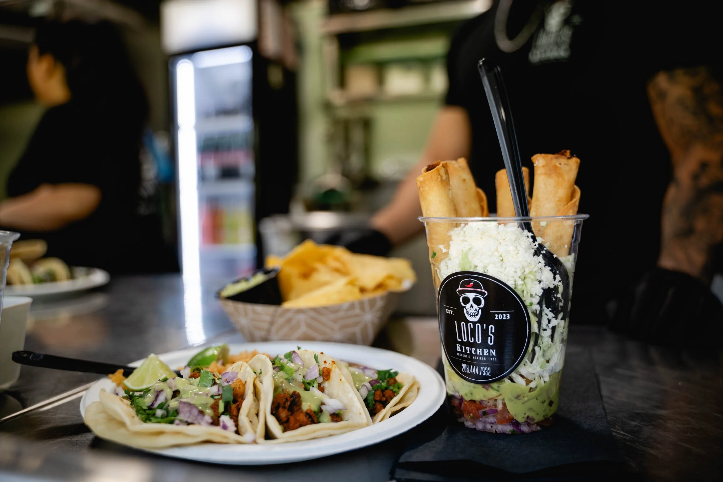 A clear plastic cup filled with chopped lettuce, shredded cheese, and fried taquitos, with a black straw, placed on a table in a restaurant. In front of the cup is a plate of street tacos with meat, chopped onions, cilantro, and lime wedges. There's also a basket of tortilla chips in the background.