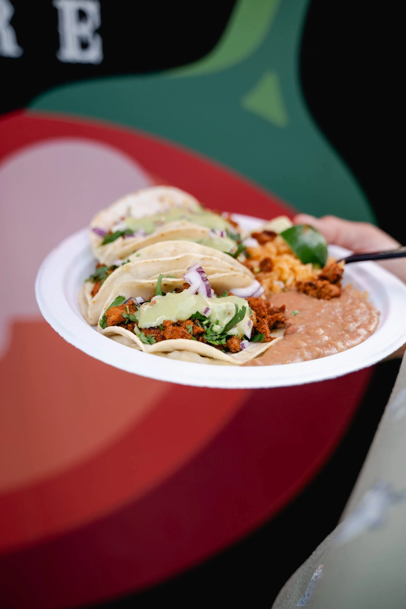 A white plate with three tacos from Loco's Kitchen topped with chopped onions, cilantro, and green sauce, served alongside refried beans, held by a person with the background featuring the Target logo.