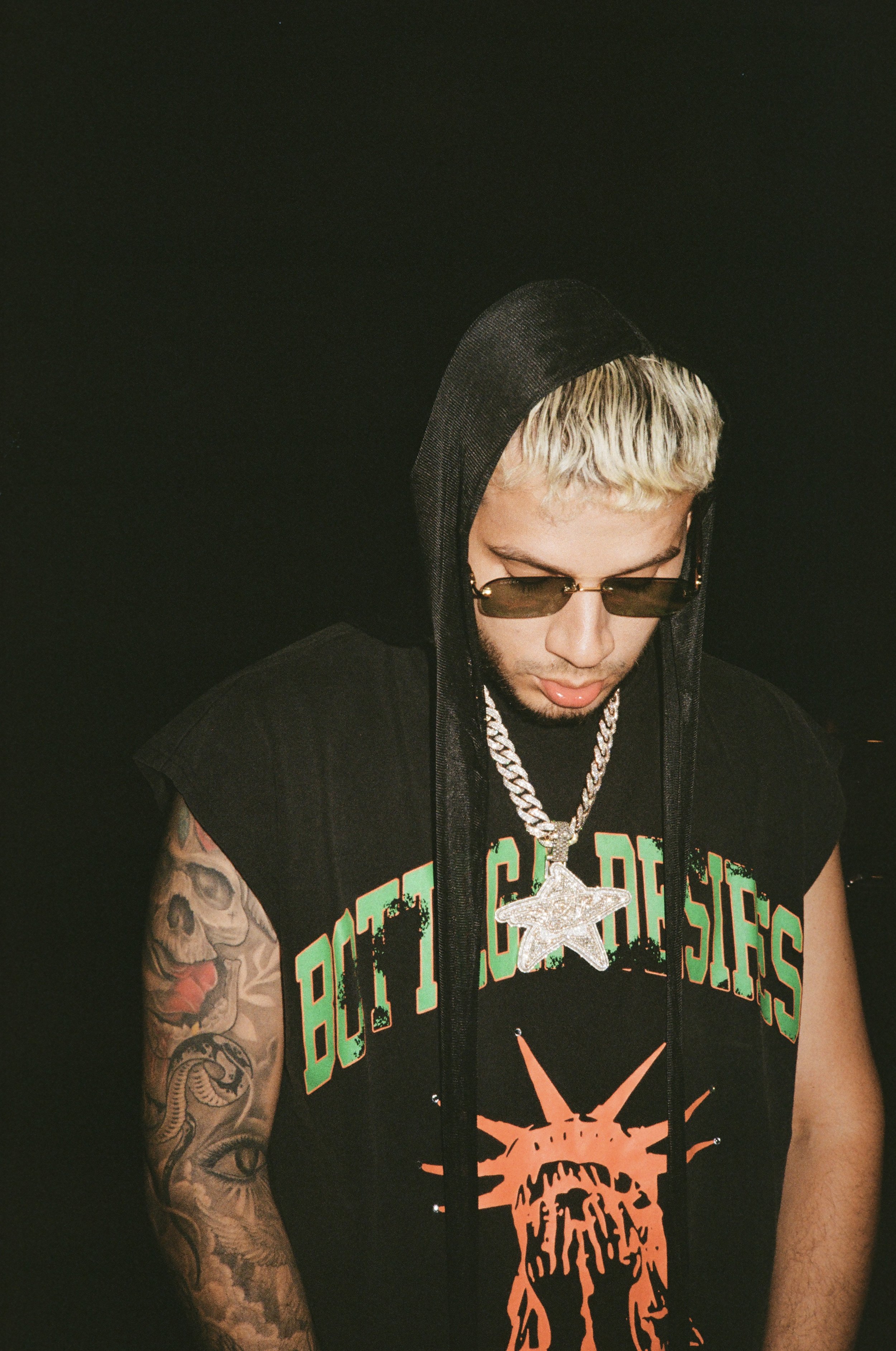 A person with bleached blonde hair wearing sunglasses, a black hoodie with the hood up, a black T-shirt with colorful text and graphic, layered silver chains with a large star pendant, and tattoos on their left arm, standing against a dark background