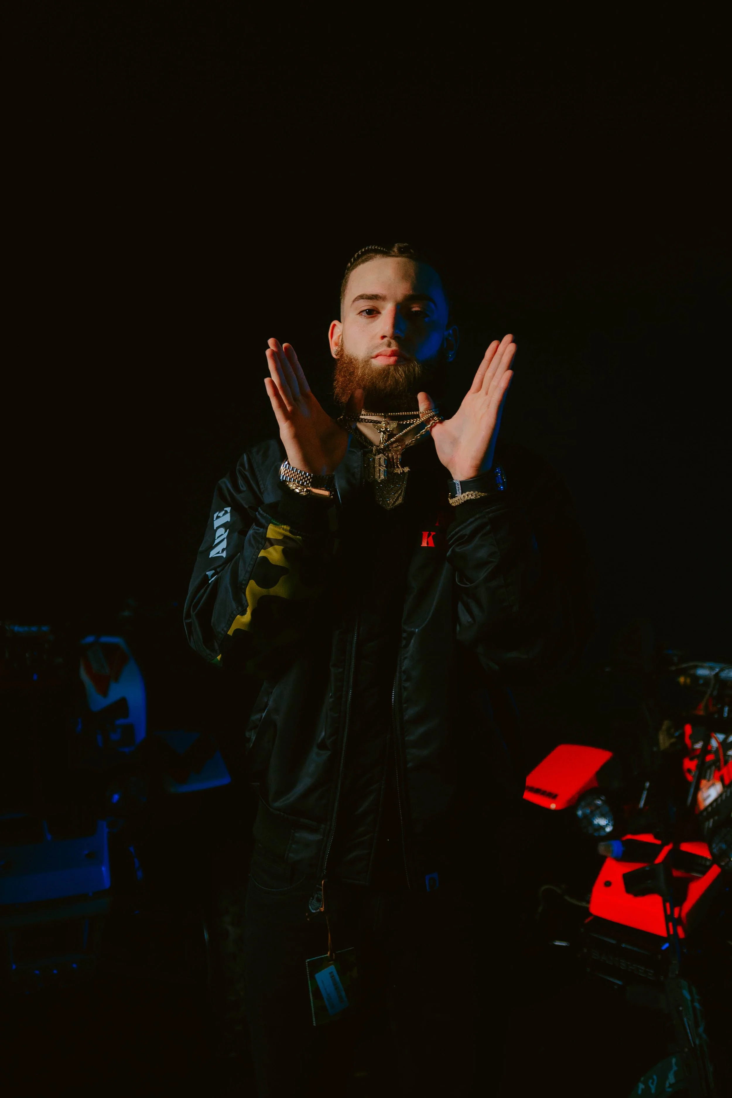 Young man with braided hair and a beard wearing jewelry and a black jacket with yellow accents, standing in a dark, neon-lit room with various electronic equipment and red lighting.