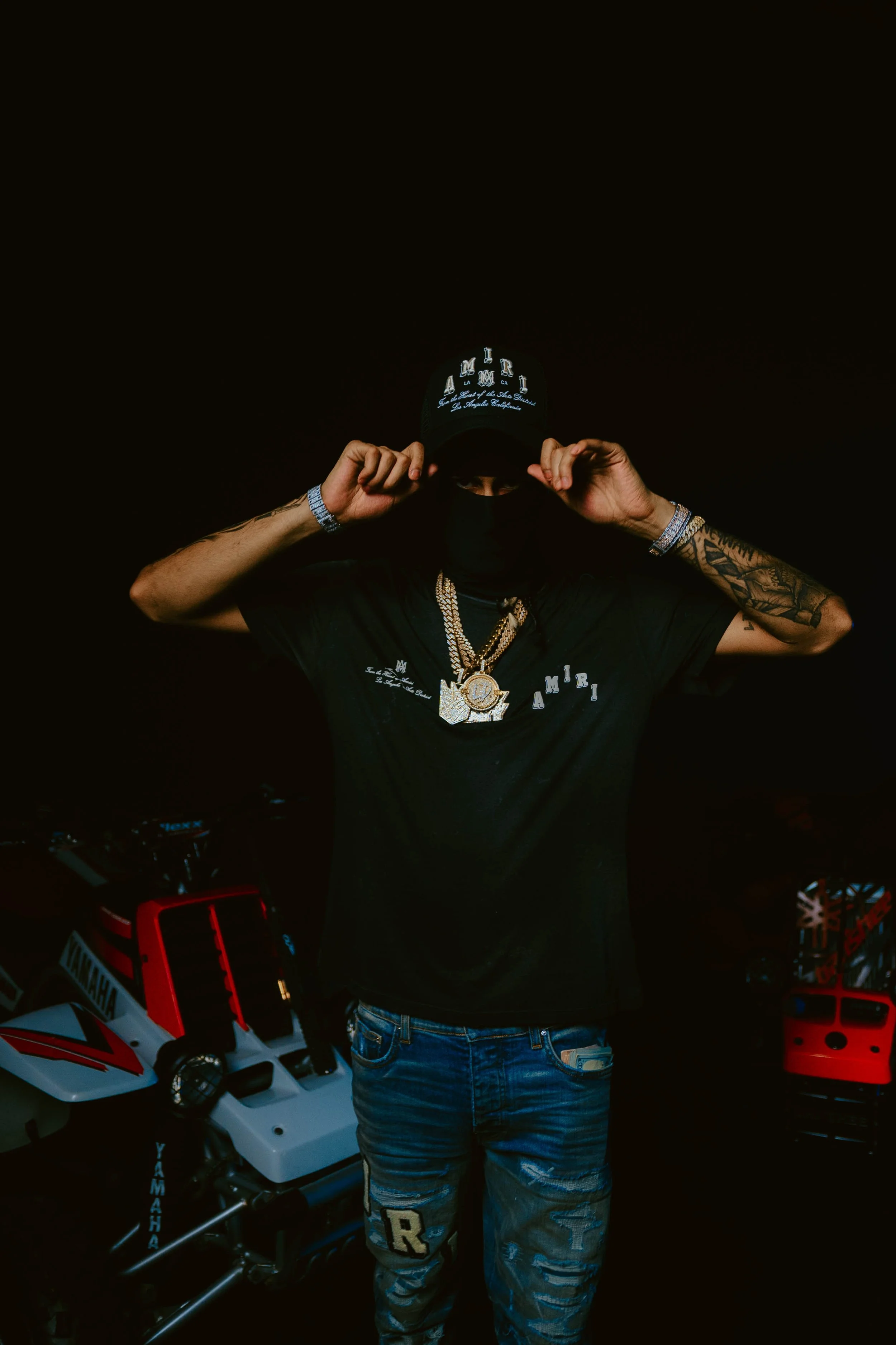 A person wearing a black baseball cap, black shirt, and jeans, holding the cap with both hands. The person has tattoos on their arms and is accessorized with jewelry. The background is dark, with some toy motorcycles visible.