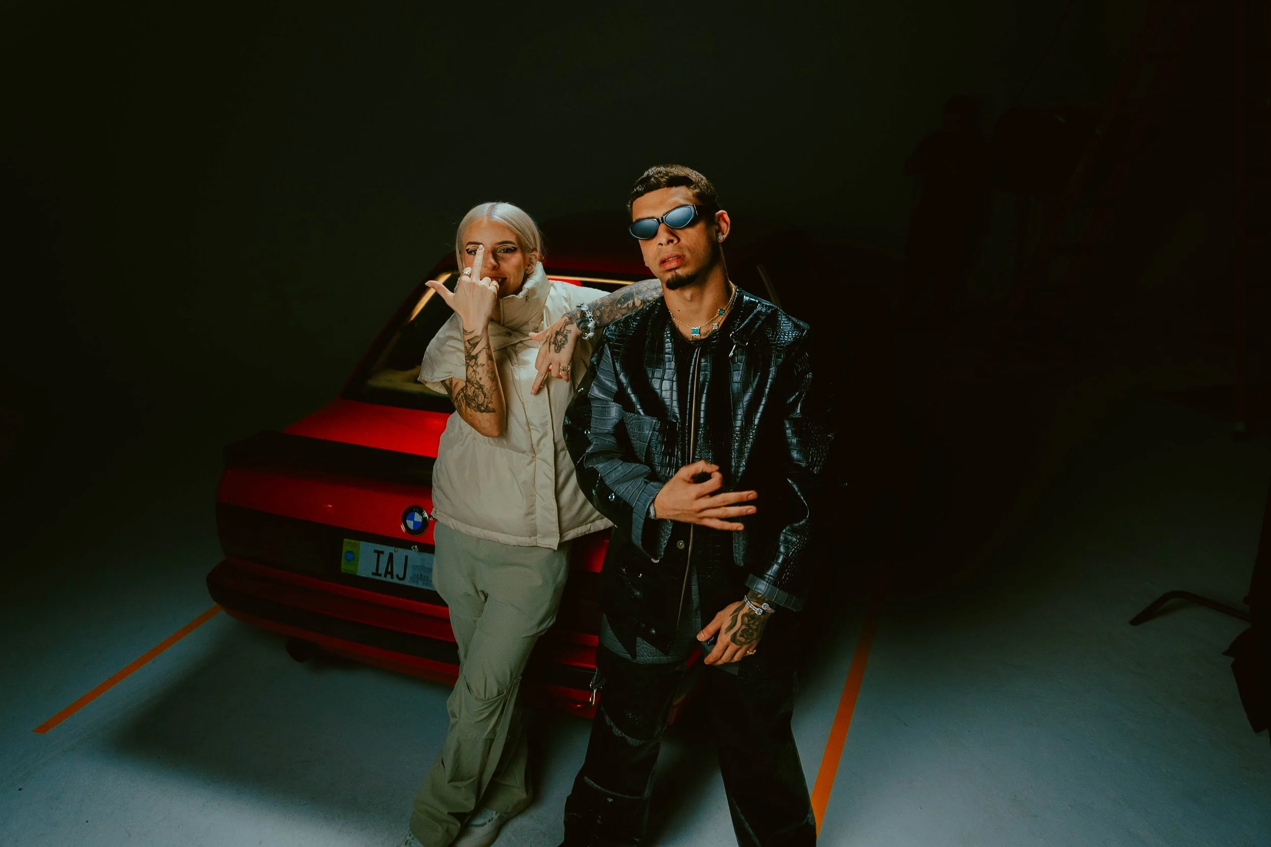 Two young adults standing in front of a red BMW car in a dark parking garage. The woman on the left has platinum blonde hair, tattoos, and is making a rock sign with her hand. The man on the right has dark hair, tattoos, and is wearing reflective sun
