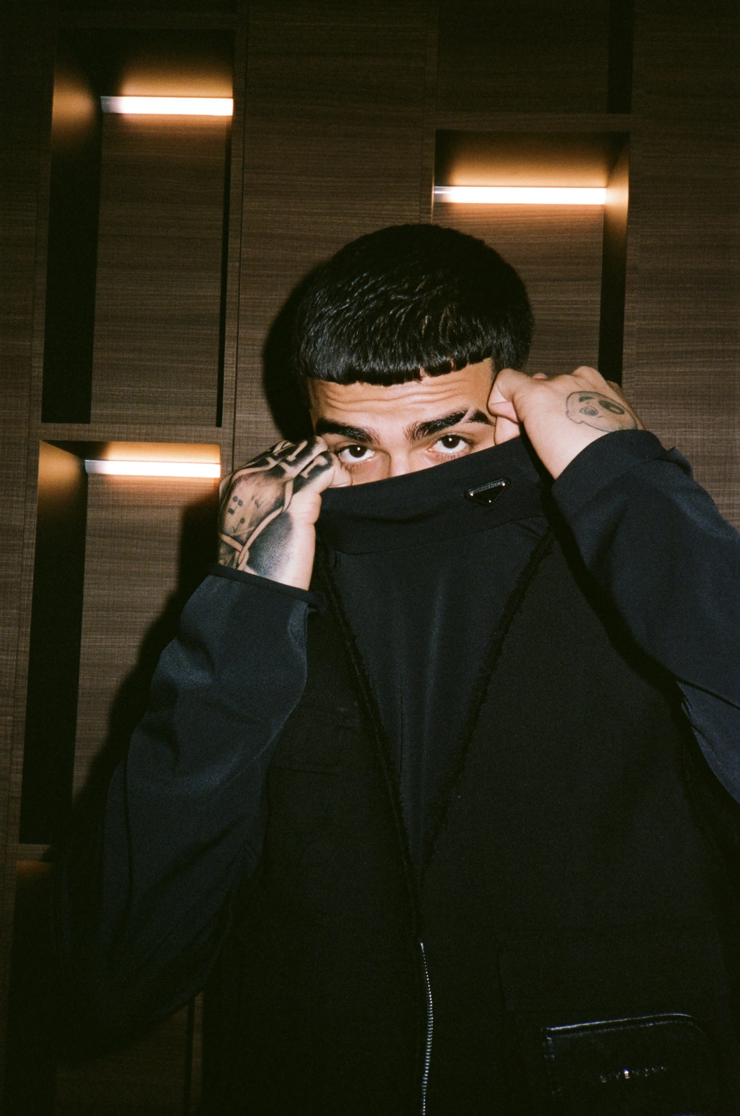 A young man with dark hair and tattoos on his hand and finger partially covering his face with a black hoodie, standing in front of a dimly lit wooden wall with rectangular light fixtures.