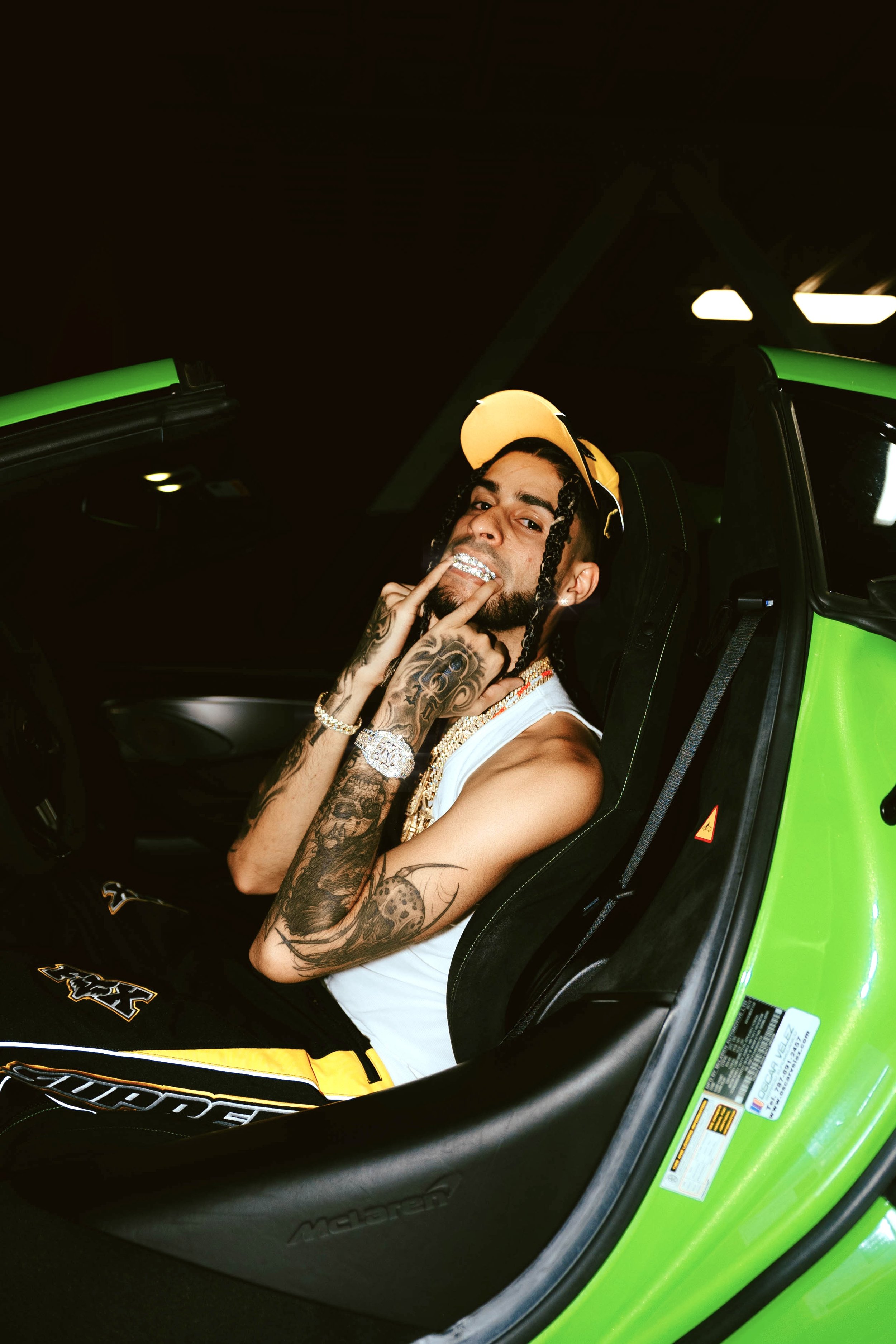 Man with tattoos wearing a white tank top, with dalmatian tattoos on his arm, sitting inside a green Lamborghini with a yellow cap, flashing a grin, showing grillz, in a dark setting with green accents.
