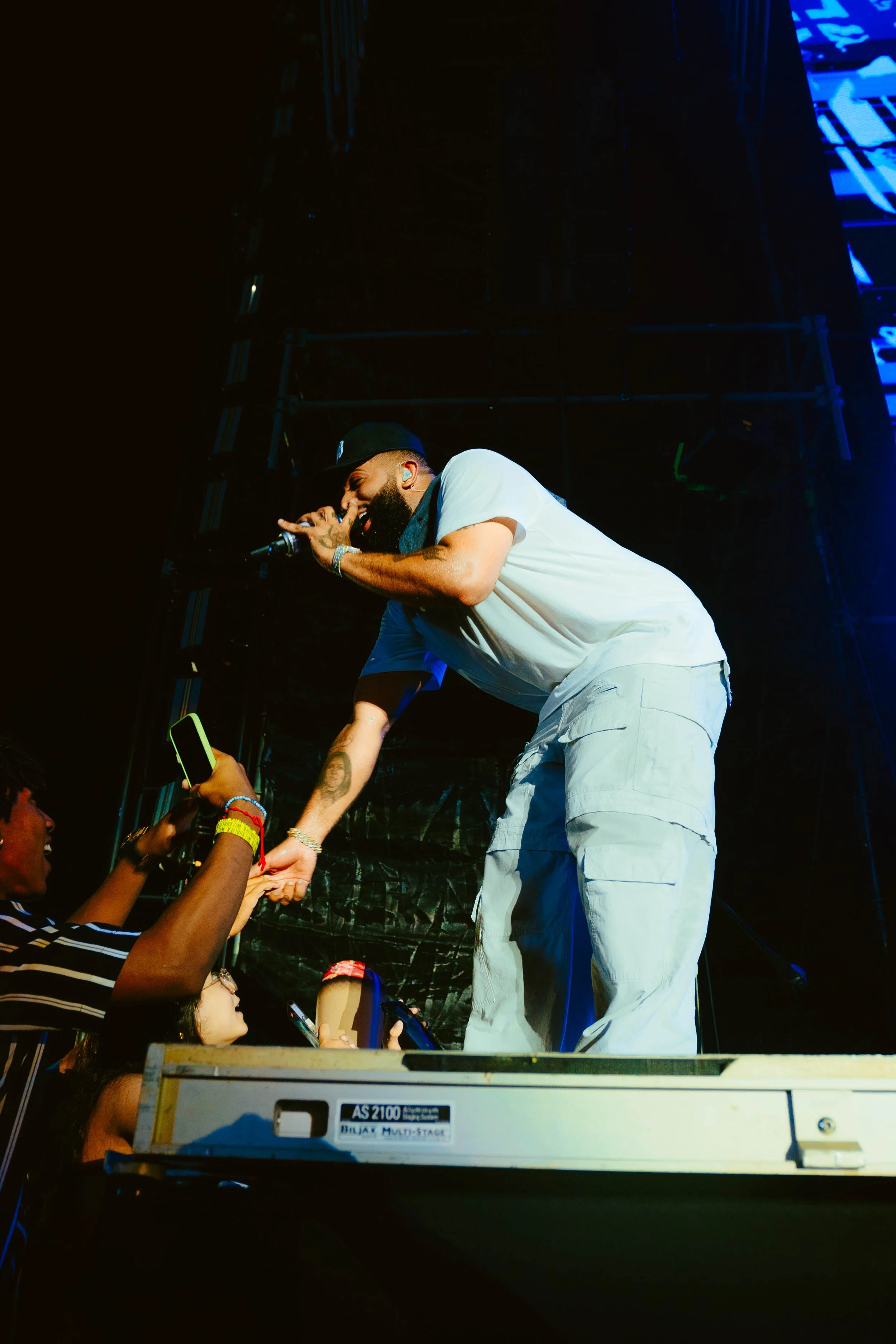 Performer on stage interacting with fans at a concert, holding hands with fans reaching out from the audience, with stage equipment visible.