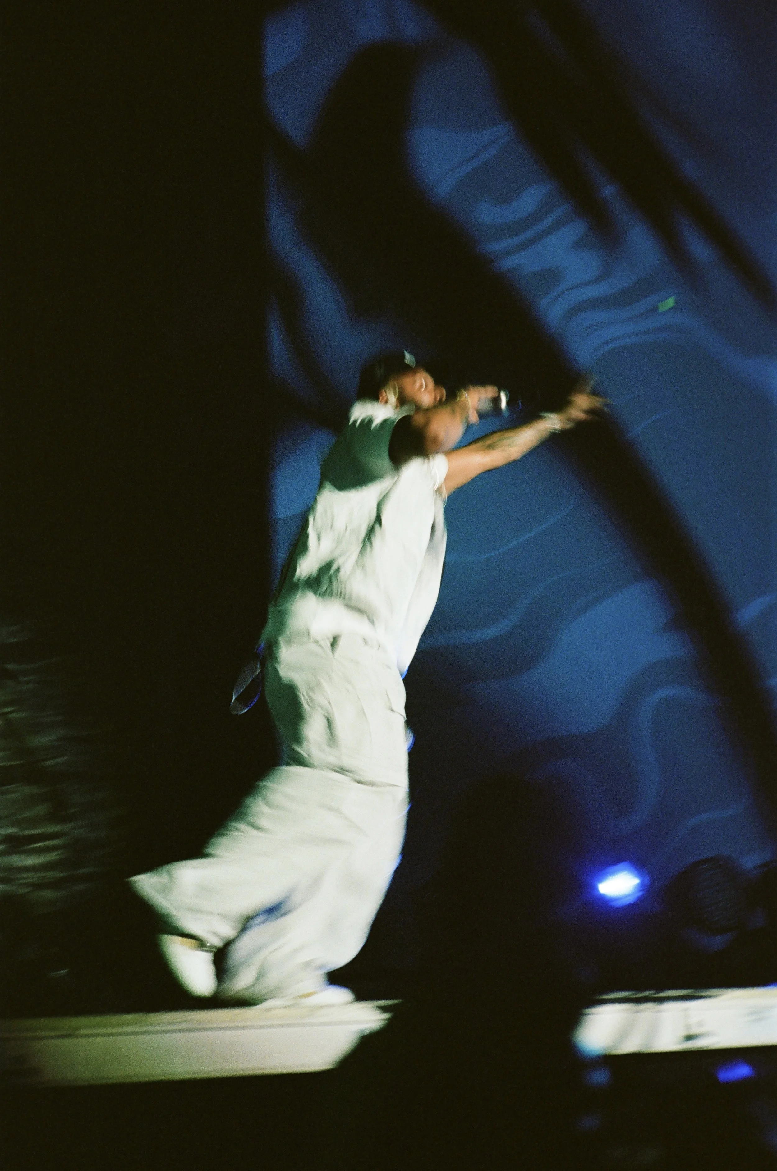 A performer on stage wearing a white outfit, mid-motion, with a microphone, blurred motion, and blue lighting effects.