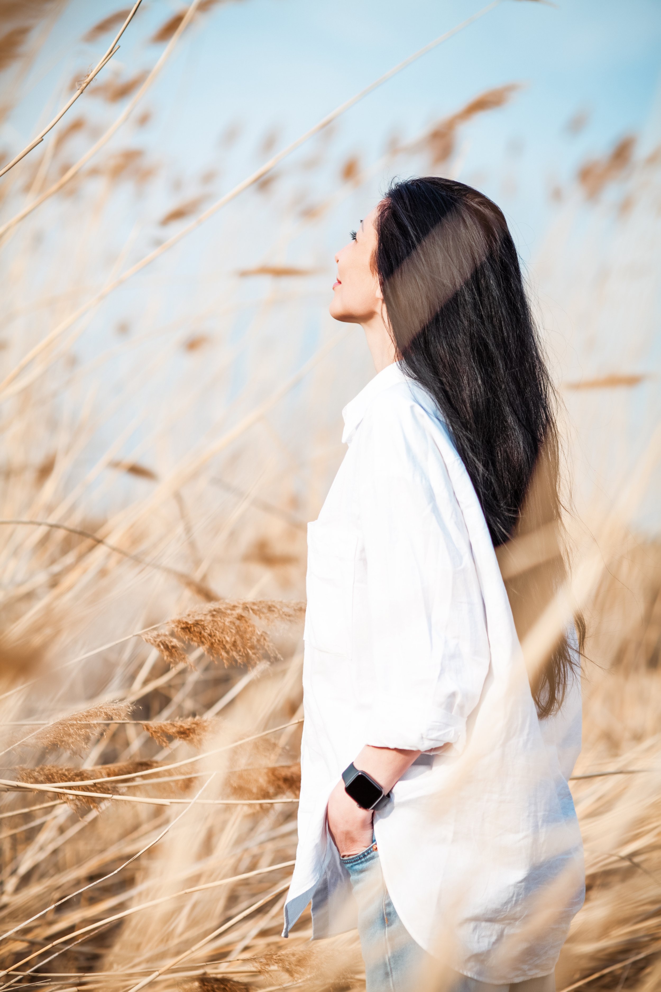 Young brunette woman with long hair, wearing white blouse, outdoors, standing in a field in wind, on blue sky background, trauma therapist near me
