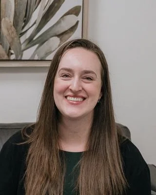 A young woman with long brown hair smiling at the camera, with window blinds in the background.