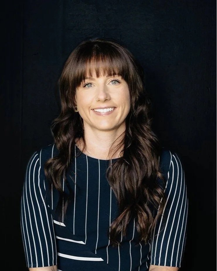 A woman with long wavy brown hair, wearing a black top, is sitting on a beige and brown upholstered chair, smiling at the camera in a well-lit room with a plain light gray wall background.