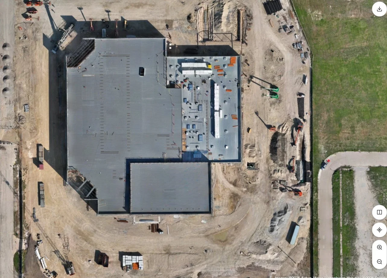 An aerial view of a building under construction with a metal roof, construction equipment, and surrounding dirt and grassy areas.