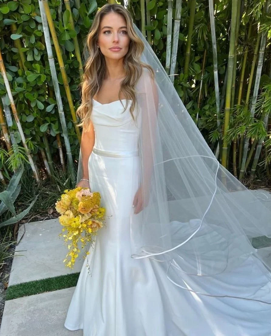 A woman in a white wedding dress holding a yellow flower bouquet, standing outdoors with green foliage in the background.