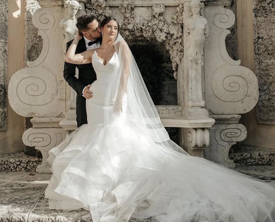 A bride and groom in wedding attire standing in front of an ornate stone backdrop with sculptures and carvings.