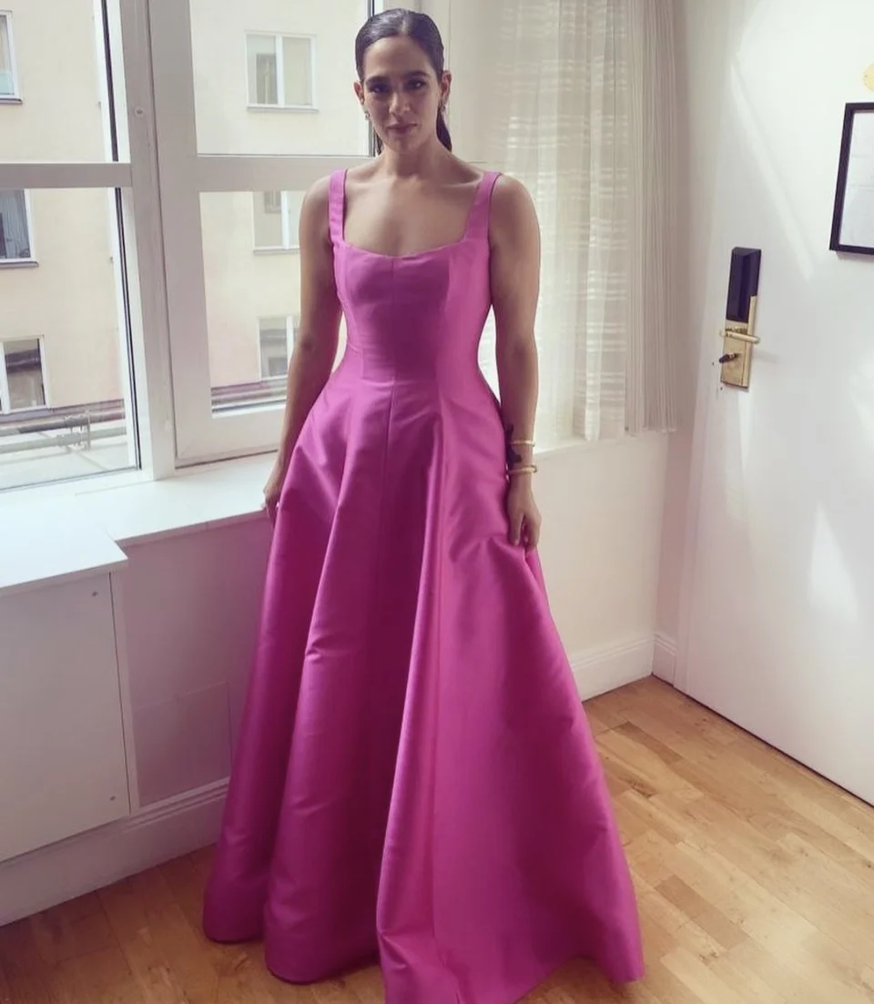 A woman is standing in a room wearing a bright pink, sleeveless, full-length gown with a fitted bodice and a full skirt, near a window with curtains.