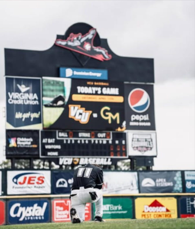 Baseball player in black jersey with number 11 kneeling on the field near the scoreboard at VCU baseball game.