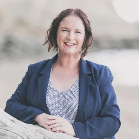 A woman with shoulder-length brown hair smiling outdoors, wearing a navy blue blazer and a patterned shirt.