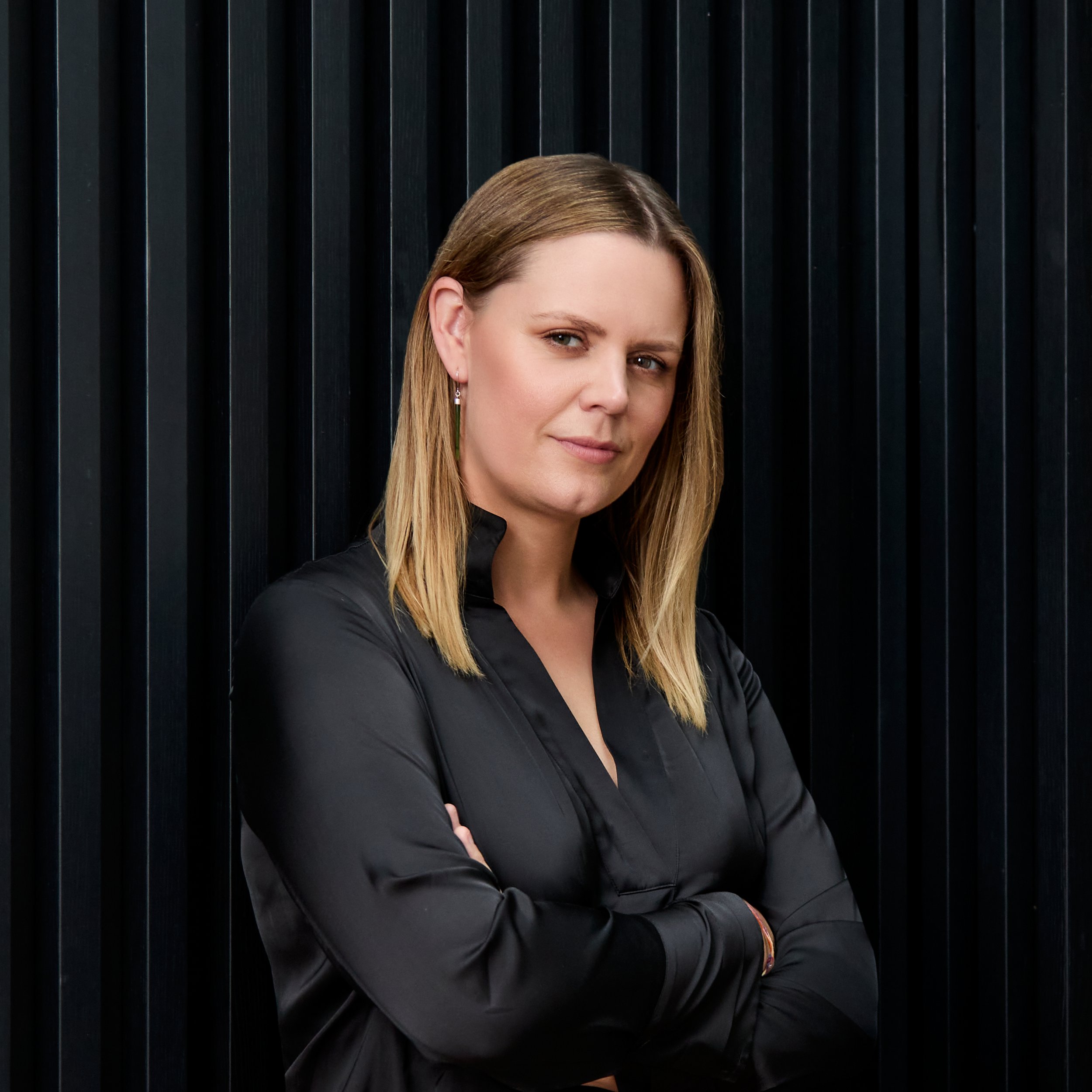 A woman with light brown hair, wearing earrings and a dark top, stands with her arms crossed in front of a black vertical striped background.
