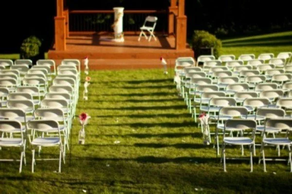 Empty outdoor wedding ceremony setup with chairs on grass, decorated with flowers, facing a decorated wooden platform with a chair and columns, in sunlight.