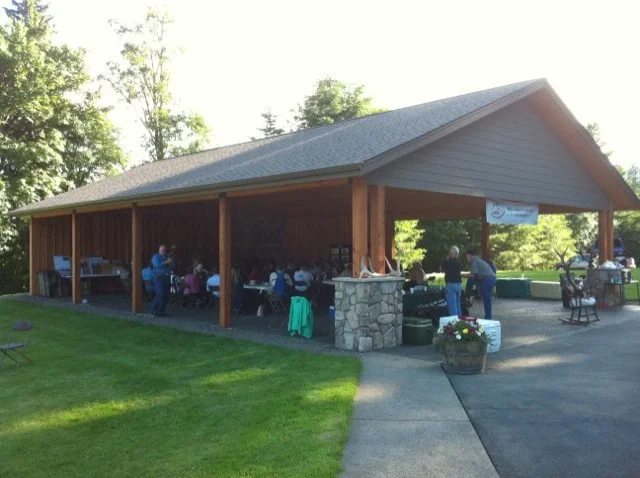 A gathering of people under a large wooden pavilion with a gabled roof in a park-like setting, with grass, trees, and a stone column at the entrance.