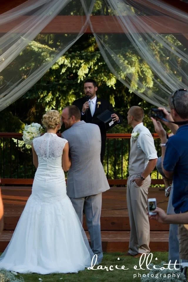 A wedding ceremony taking place outdoors under a wooden pergola decorated with sheer white curtains. The bride in a white wedding gown and the groom in a light gray suit are standing in front of a man in a black suit who appears to be officiating, ho