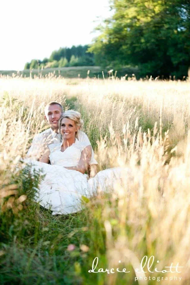 A couple sitting together in a field of tall grass on a sunny day, with trees in the background.