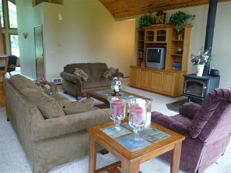 Living room with three sofas, a wooden coffee table, and an entertainment center with a TV. There's a wood stove with a vase of flowers next to it and a small side table with candles.